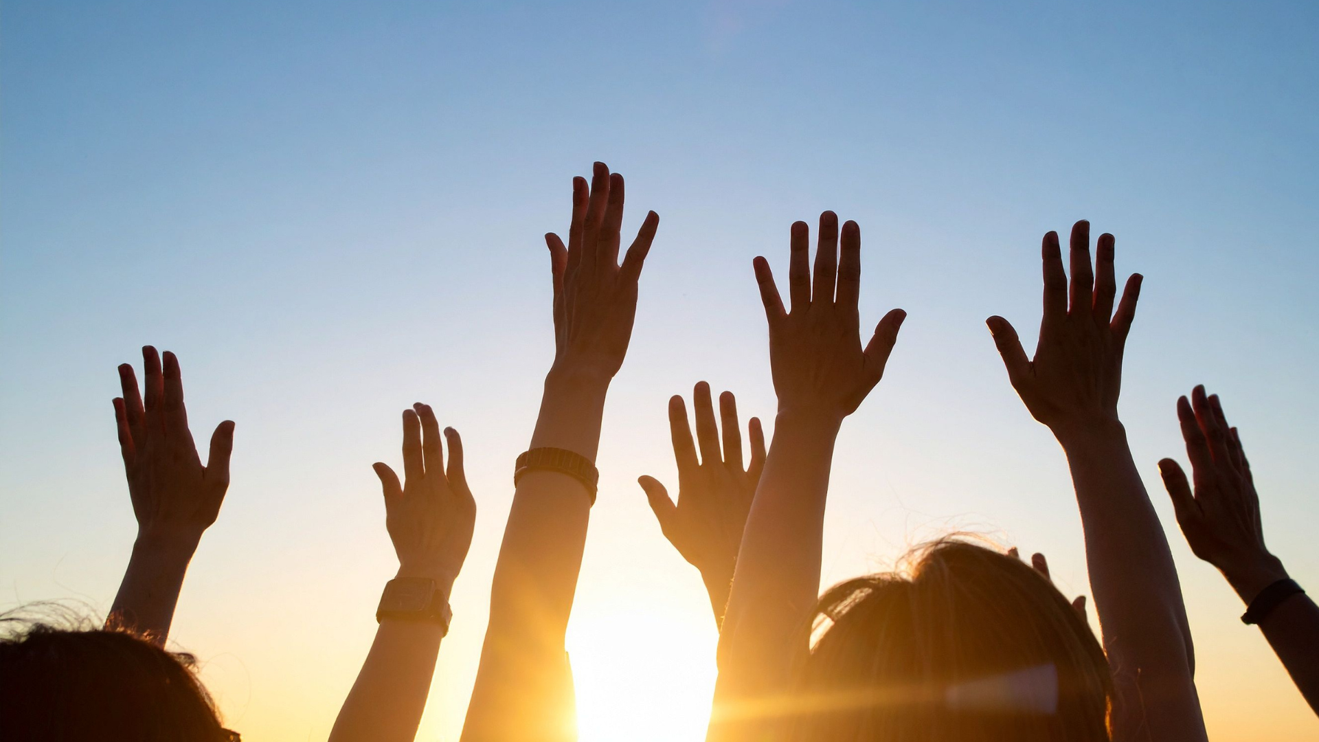 A group of people raising their hands up towards the setting sun.